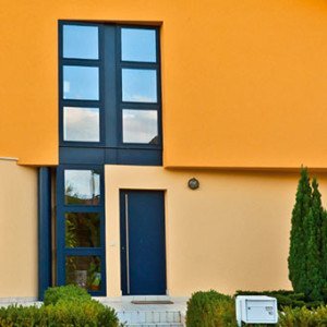 Modernes Haus mit blauer Tür und großen Fenstern, umgeben von grünen Pflanzen und einem gelben Fassa.