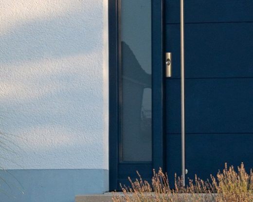 Blick auf moderne Haustür mit blauer Fassade und Gartenpflanzen.