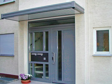Hauseingang mit moderner Tür und großen Fenstern, umgeben von Bäumen und Sträuchern.