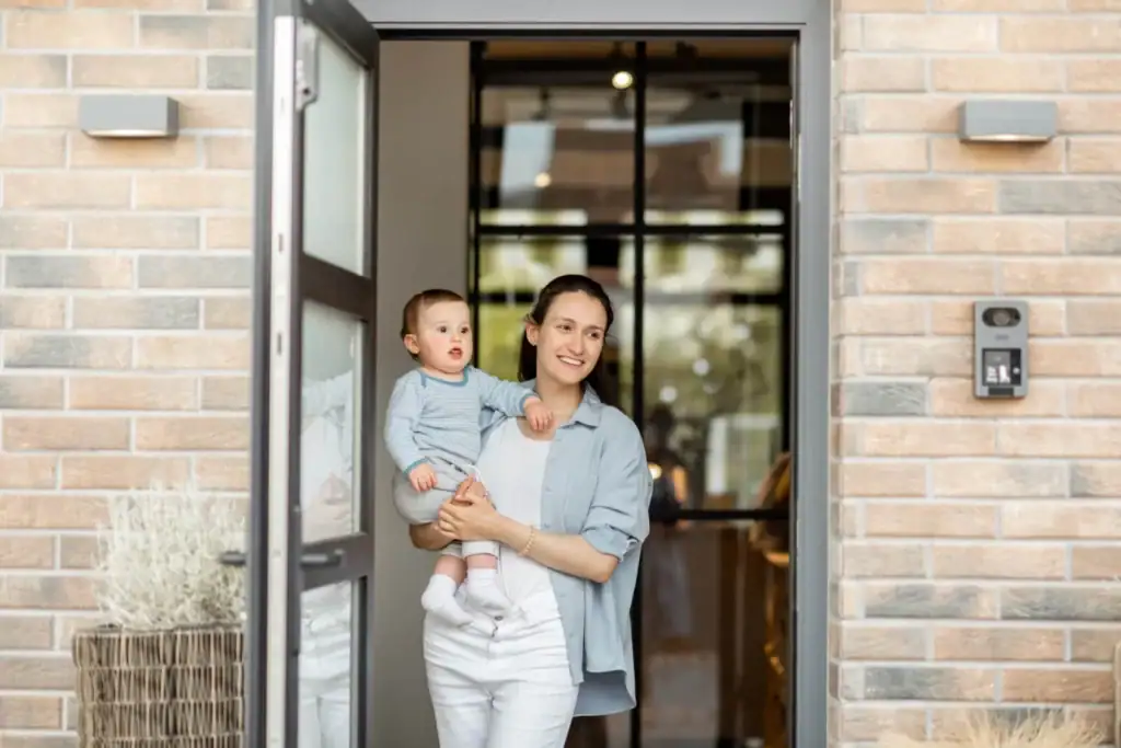 Fröhliche Mutter mit Baby am Hauseingang, moderne Haustür mit Sicherheitskamera, Wohnkomfort.