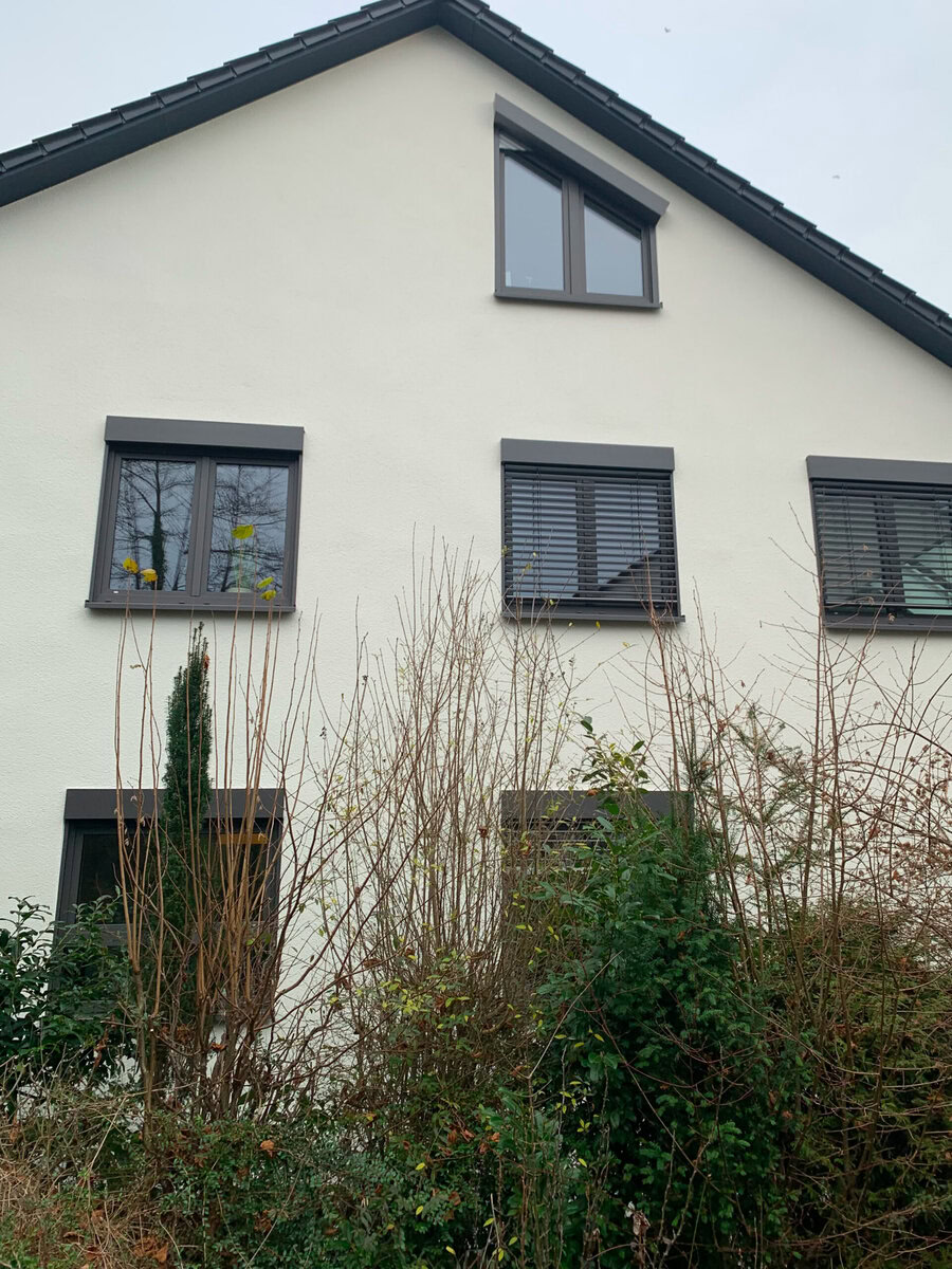 Moderner Mehrfamilienhaus mit dunklen Fensterrahmen und Außenjalousien, umgeben von winterlichem Bewuchs.